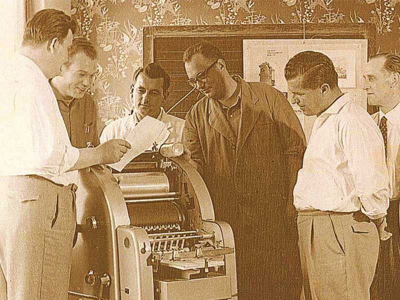 Sechziger Jahre, sechs Geschäftsmänner bestaunen historischen Kopierautomat in der Mitte des Bildes, 