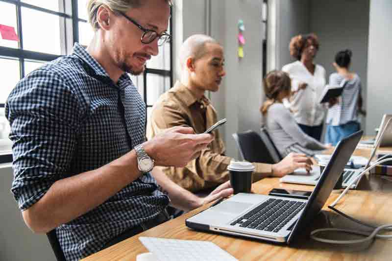 Arbeitsalltag im Büro, zwei Männer vor Laptop arbeiten, Mann hat Handy in der Hand, Hintergrund  3 Personen im Gespräch