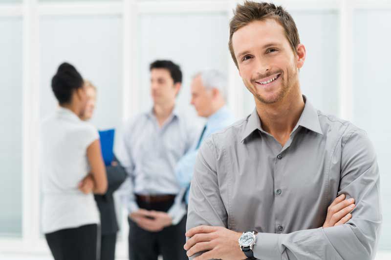 sympathischer Geschäftsmann schaut direkt den Betrachter an, lächelt, graues Hemd, moderne Uhr, im Hintergrund Businessmeeting vier Personen im Gespräch