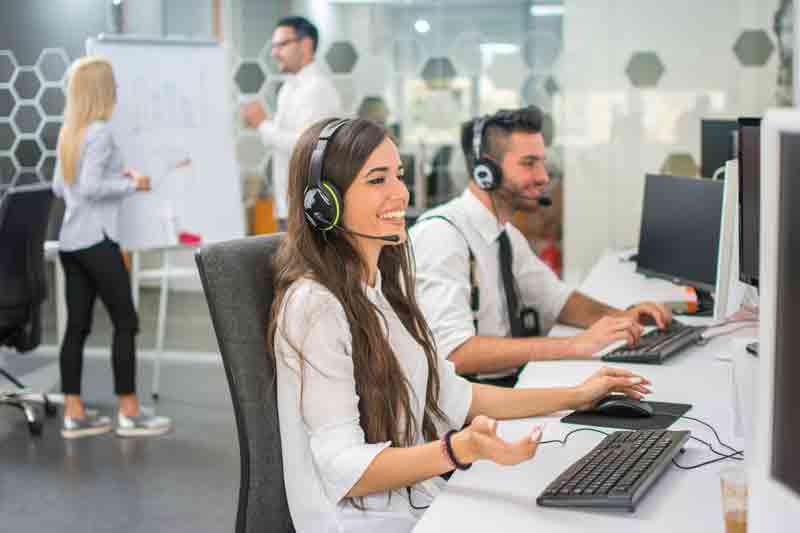 Frau und Mann mit Headset telefonieren vor PC, im Hintergrund Gespräch vor Flipchart